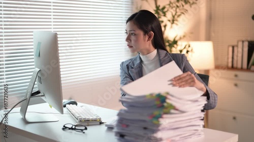 A woman is sitting at a desk with a stack of papers in front of her. She is looking at the papers and she is working