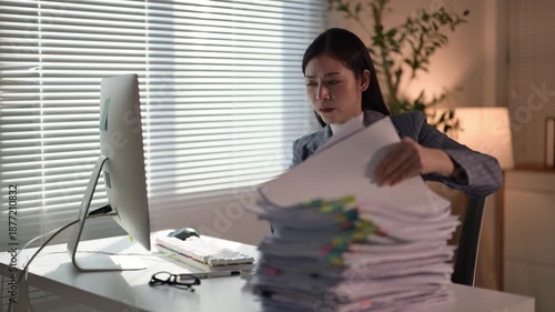 A woman is sitting at a desk with a stack of papers in front of her. She is looking at the papers and possibly reading them