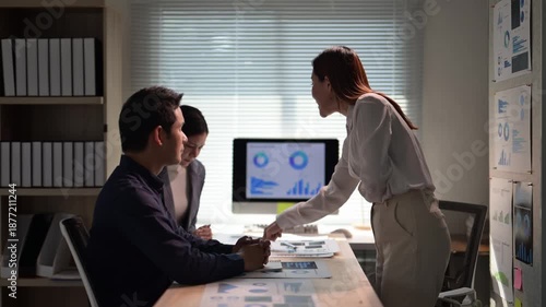 A woman stands in front of a group of people in a conference room. There are two monitors on the table in front of the group