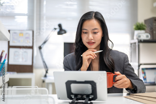Asian businesswoman or office worker with a pen under chin holding coffee looking at tablet on table