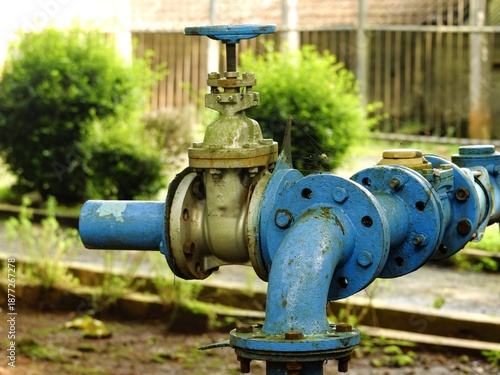 Close-up of an industrial blue gate valve and piping system outdoors. This weathered water infrastructure shows signs of moss and age, representing utility management and engineering.