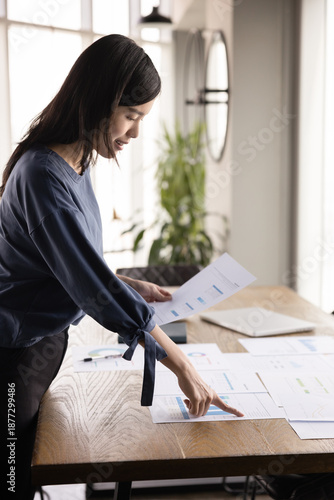 Focused busy young 20s manager woman analyzing project benchmarks, reviewing paper marketing reports with diagrams on office table, thinking on ideas for growth, smiling