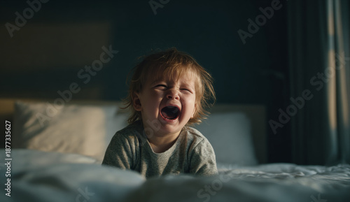 Young toddler with light brown hair crying and screaming on a bed in a dimly lit room, expressing distress or frustration