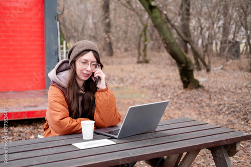 A young female freelancer in a brown jacket and hat works remotely at a table in an autumn park, making a business call. Conceptual concepts of freelancing, online business