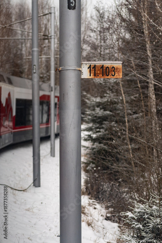 A close-up shot of a gray metal railway pole with a numbered orange sign, set against a background of a snowy forest and a red and white passenger train on the tracks.