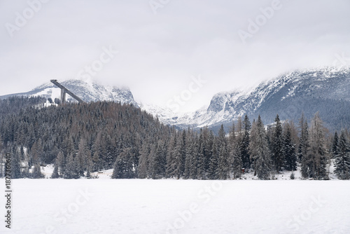 Wide panoramic view of a frozen white lake in the foreground with a ski jump structure nestled in a dense pine forest against snow-covered mountain peaks and a misty sky.