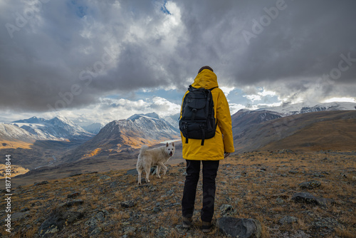 Tableau sur toile A woman in a yellow jacket and black backpack stands on a mountain road rise, gazing at a valley with snow-dusted peaks and colorful forest