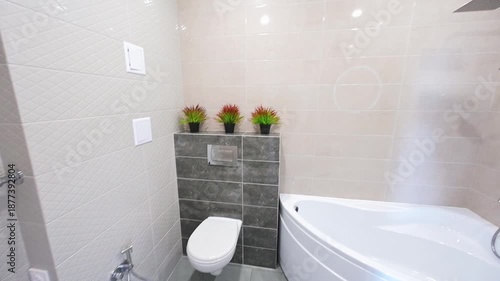 modern bathroom with a white bathtub, wall-mounted shower system, round mirror, light beige tiles, dark grey accent tiles, three small potted plants on a ledge.