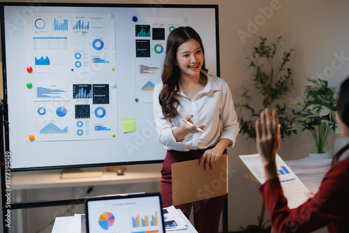 Young Asian businesswoman smiling while presenting financial charts and data on a whiteboard, a colleague raising hand asking a question during a corporate meeting in an office setting
