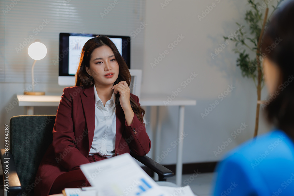 Obraz premium Asian businesswoman in a maroon suit attentively listening to a colleague or client, analyzing financial documents during a focused business meeting in a modern office setup