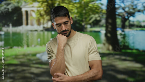 Pensive man outdoors in park setting with thoughtful expression wearing casual beige shirt surrounded by lush trees and peaceful lake under vibrant sunlight