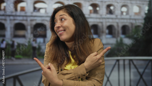 Bilde på lerret Smiling hispanic woman pointing fingers to sides at sunlit ancient roman coliseum building; amusement