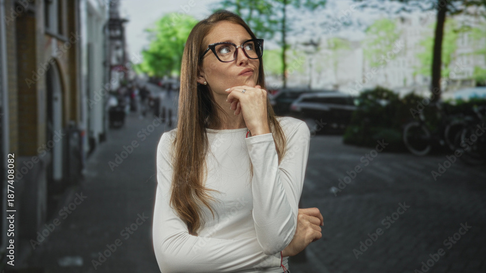 Fototapeta premium Woman in white top wearing glasses with hand on chin and crossed arms on street, looking up and pondering; pensive doubt decision.