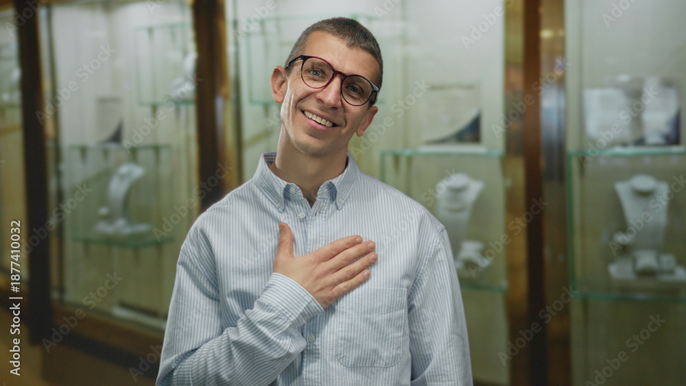 Obraz premium Man smiling indoors near jewelry showcase wearing glasses and striped shirt with one hand on chest in a store setting expressing satisfaction and positive emotion.