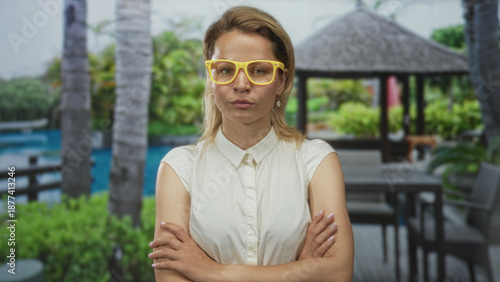 Woman wearing yellow glasses crosses arms on a hotel terrace beside a pool at a resort building; rejection.