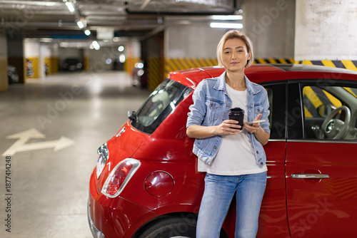 A woman stands next to her red car in an underground parking garage, holding a coffee and her phone