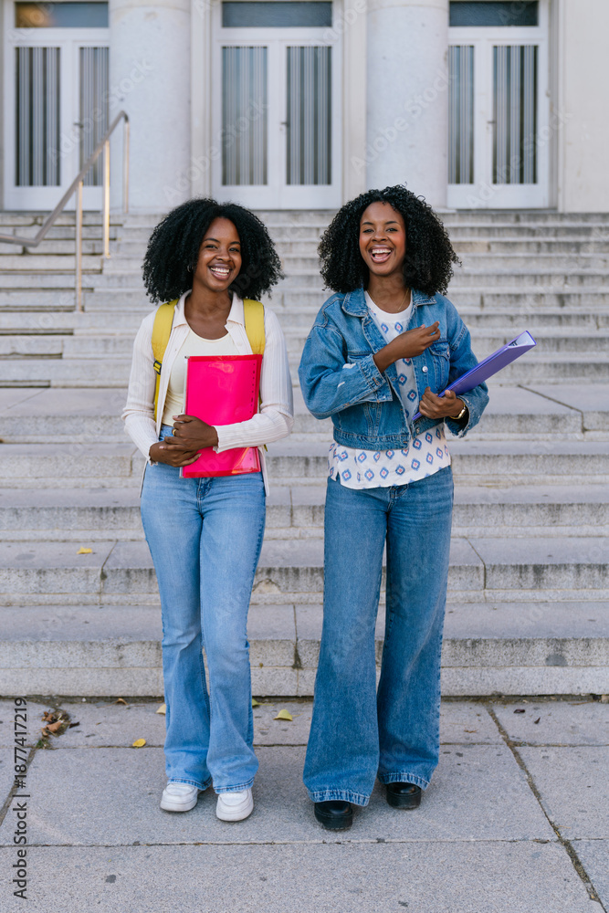 Fototapeta premium Two happy female students holding books and smiling while walking down stairs on a university campus