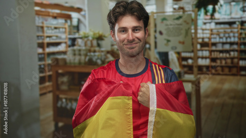 Man wearing red soccer jersey holding a spanish flag and clenching fists in a shop interior while smiling; pride support celebration.