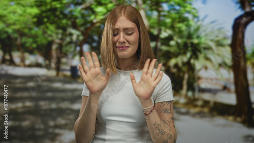 Obraz na plátne Young blonde woman holds up bare palms with wrinkled nose and pursed lips in disgust gesture at sunny park pathway; disgust repulsion aversion distaste