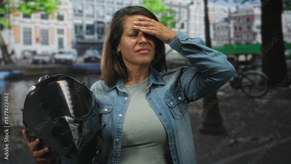 Fototapeta premium Middle age hispanic woman holding glossy black motorbike helmet, hand pressed to forehead for headache beside canal street with parked bicycles; exhaustion.