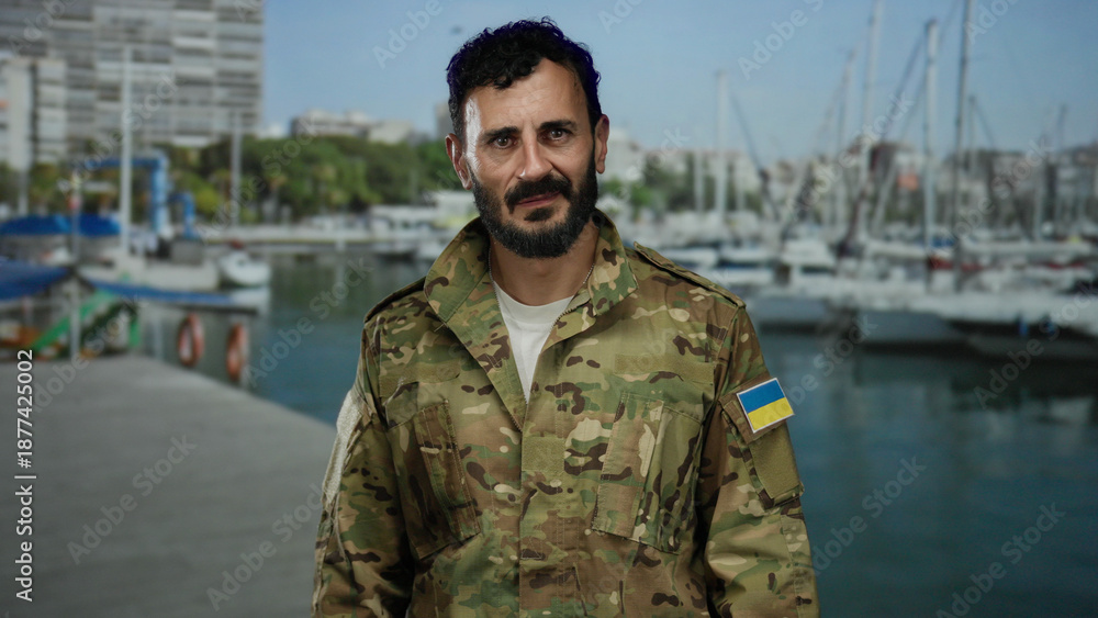 Fototapeta premium Hispanic man in military uniform with ukrainian flag patch stands near port with boats and ships, showcasing a maritime military scene with a calm sea backdrop.