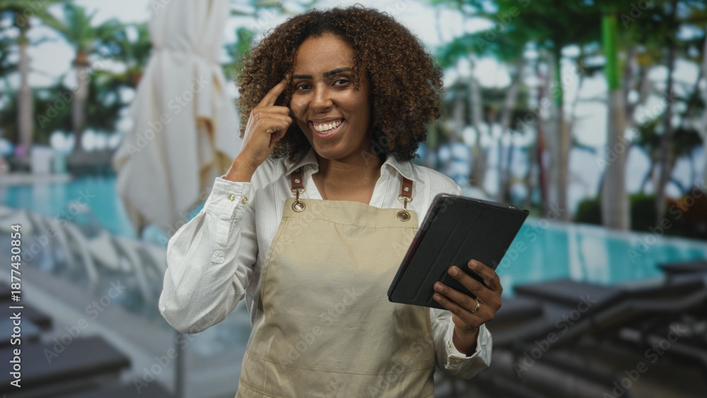 Fototapeta premium Woman in apron points finger to temple while holding tablet in building pool area, smiling and checking notes on device; confidence hospitality.