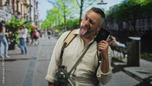 Quadro su tela Man middle aged winking and sticking out tongue holding passport on city street while wearing backpack and canteen; playful wanderlust