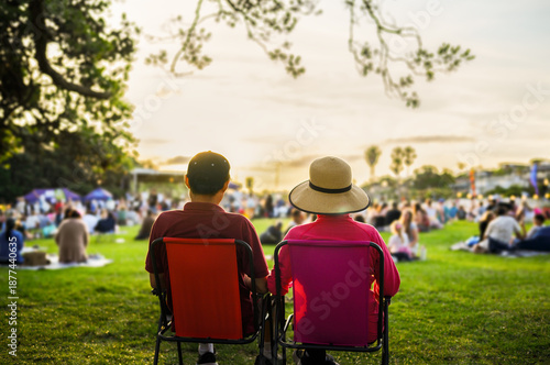 Couple sitting on the picnic chairs, listening to the music concert. Unrecognizable crowd of people in the park.
