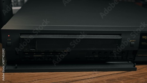 Male hand presses eject button on black vintage VCR player to release cassette. Person removes analog tape from retro video recorder on wooden surface.