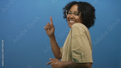 Photos Woman wearing glasses points finger and smiles in blue studio; happiness optimism confidence energy