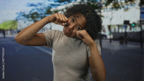 Foto Woman making tearful expression with fists pressed to cheeks, puckered lips, and scrunched eyes in a sunlit city street; melancholy