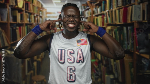 Bilde på lerret Man with fingers in ears grimacing in library wearing usa jersey and wristbands; study distraction frustration