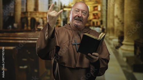 Bild auf Leinwand Senior man monk holds bible and makes rock horns gesture while smiling at camera inside ornate church building, wearing brown habit and cross necklace; playful devotion