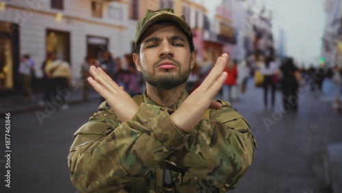 фотографія Young hispanic man in camouflage uniform makes arms crossed x gesture on crowded downtown city street; denial