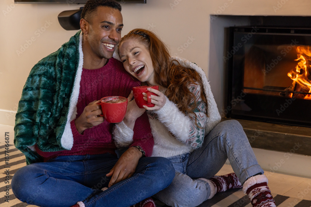 Obraz premium Diverse couple sitting in living room wrapped in green fleece blanket with red mugs by fireplace