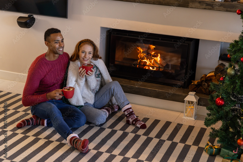 Fototapeta premium Diverse couple sitting on striped rug in front of fireplace by christmas tree with red mugs