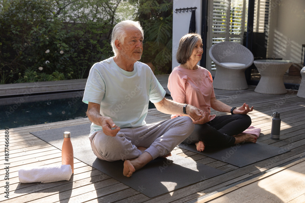 Fototapeta premium Senior couple meditating at backyard deck beside pool with yoga mats, towel and water bottles