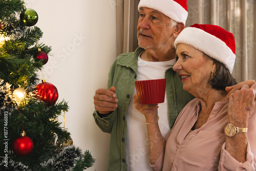 Senior couple wearing Santa hats standing next to decorated Christmas tree at home, holding red mug