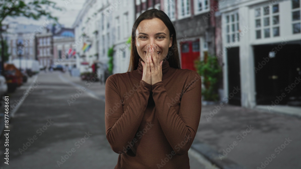 Fototapeta premium Woman with hands clasped near face smiling broadly while wearing brown turtleneck on a street lined with buildings; simple joy.