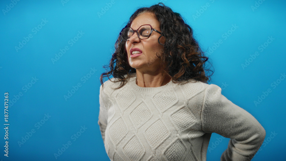 Obraz premium Woman experiencing pain, wearing glasses and sweater, standing against bright blue background, expressing discomfort and maturity, with curly hair and hispanic features.