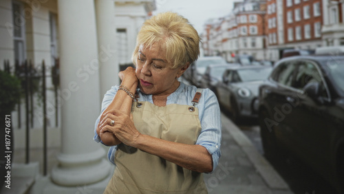 Foto Woman in apron rubs her arm and holds shoulder on street near parked car and building column; discomfort resilience