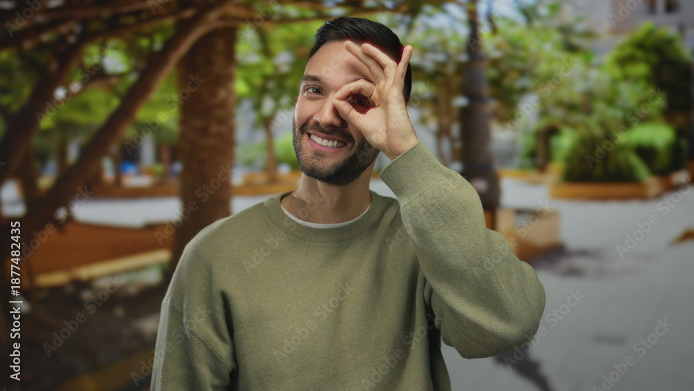 Fototapeta premium Hispanic man gesturing ok sign in a sunny park, surrounded by green outdoor scenery, expressing joyful playfulness with nature in the background.