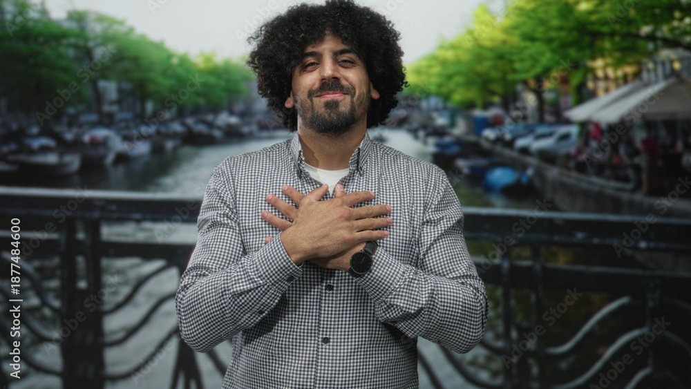 Fototapeta premium Man with curly hair and hands crossed on chest on street in amsterdam near canal bridge, smiling gently; gratitude.