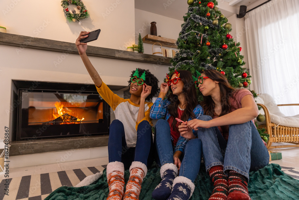 Fototapeta premium Female friends sitting on rug next to fireplace holding smartphone taking selfie by christmas tree
