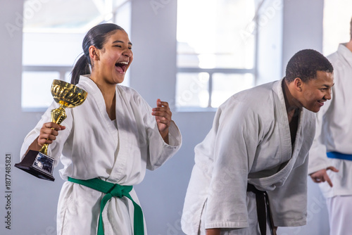 African american teen in gi holding golden trophy, laughing while instructor bowing in dojo