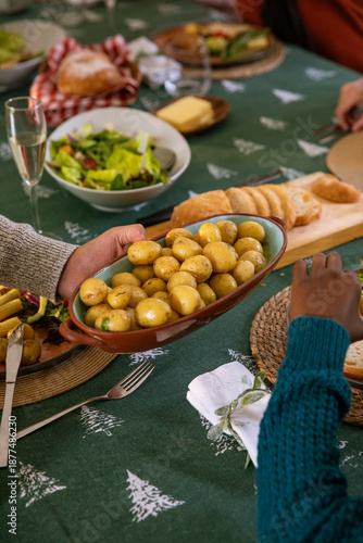 Diverse friends in sweaters passing brown ceramic dish of potatoes at home dining table