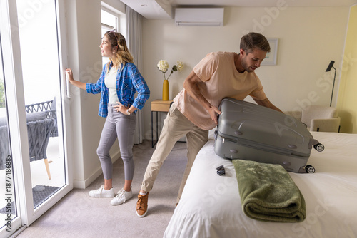 Couple lifting gray hard-shell suitcase onto bed and opening glass door to balcony in hotel room