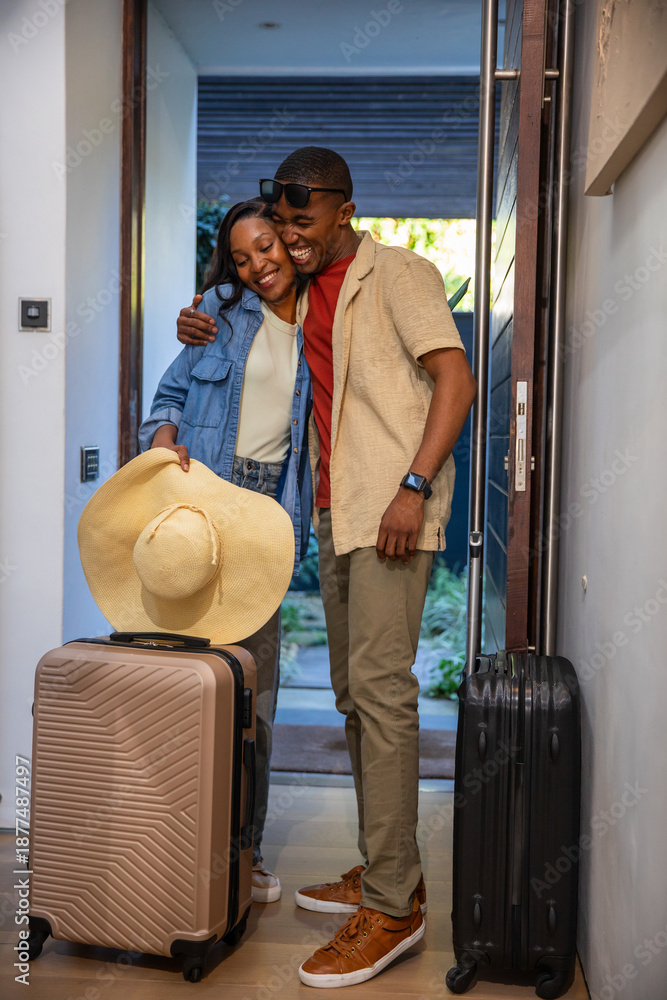 Fototapeta premium African American couple embracing in entryway by front door, holding sunhat near suitcases
