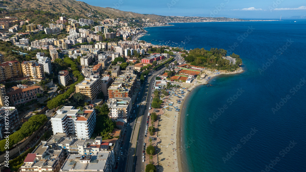Fototapeta premium Aerial view of Messina seafront, Sicily, Italy. Panorama of a coastal city nestled between green hills and the blue sea. A long sandy beach borders the urban landscape under a clear Mediterranean sky.