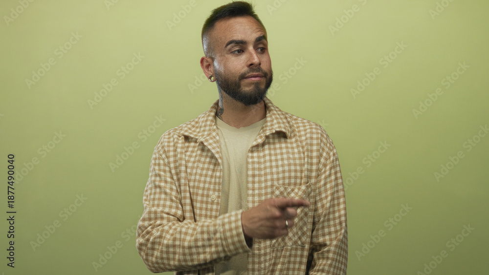 Fototapeta premium Man pointing finger in studio against olive green backdrop wearing checked shirt and rings on hand; thoughtful curiosity.